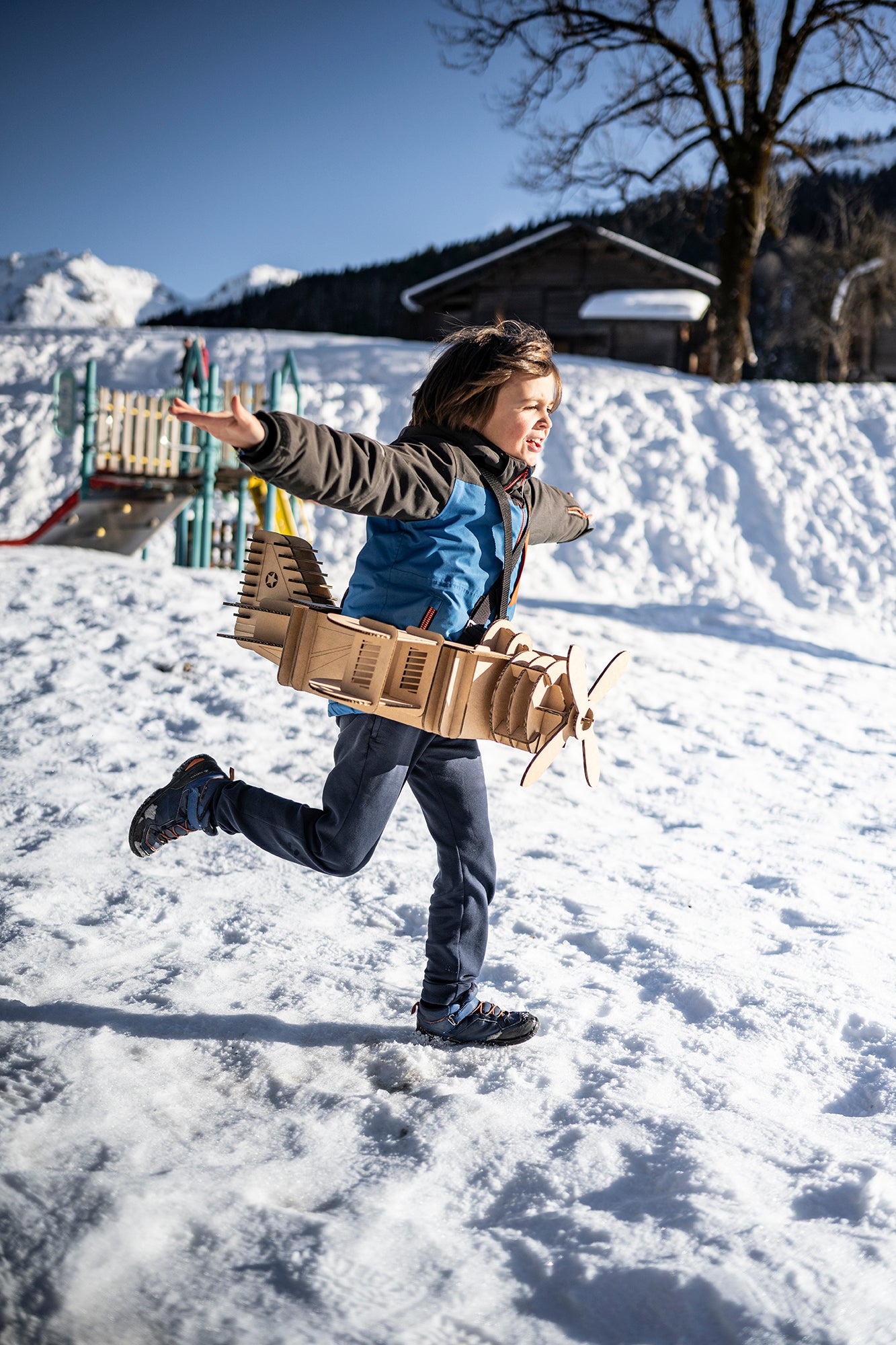 Un enfant en déguisement d'avion en carton recyclé court avec les bras en l'air dans la neige