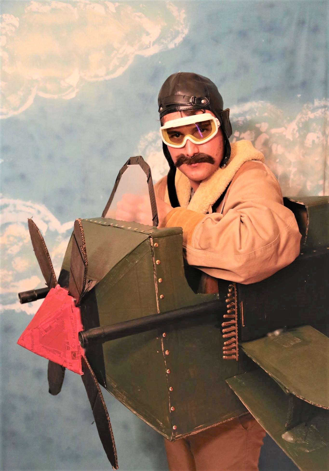 Un homme en déguisement d'avion en carton recyclé avec des mitraillettes et des lunettes sur un fond bleu à nuage