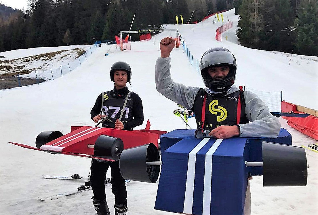 Deux hommes en déguisement de formule 1 en carton recyclé peint en rouge et bleu sur de la neige à l'arrivée du super slalom