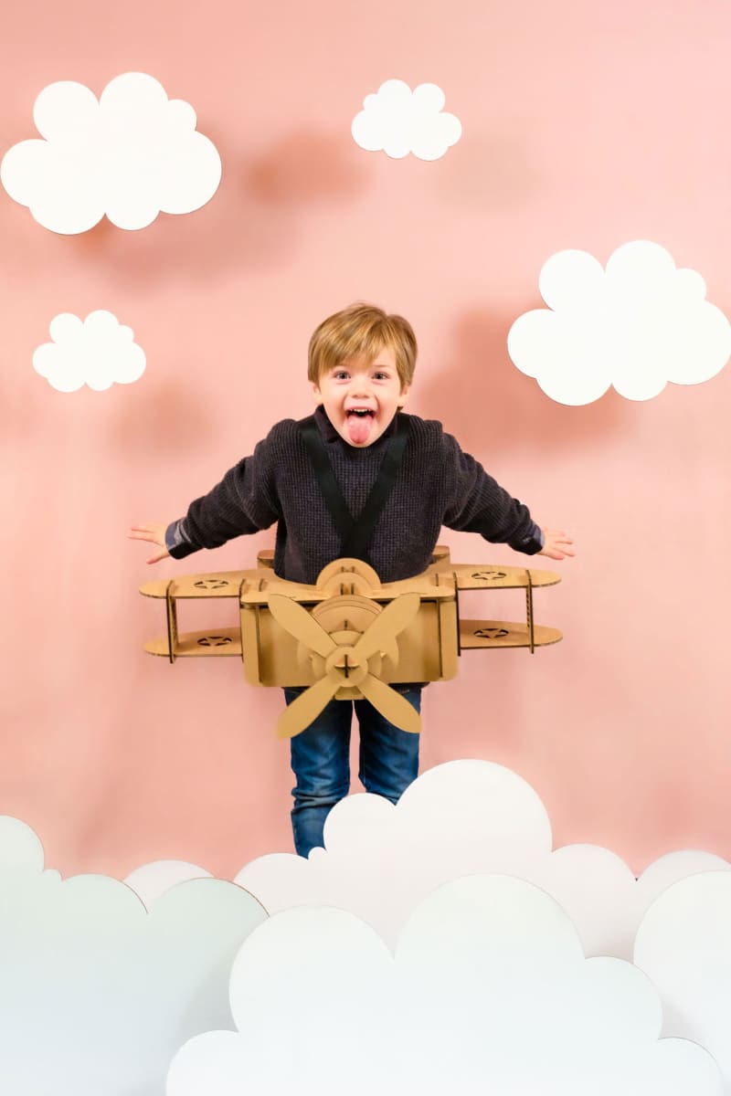 Un enfant de face qui tire la langue en déguisement d'avion en carton recyclé à construire, peindre et colorier soi même dans les nuages et un décor en carton sur fond rose