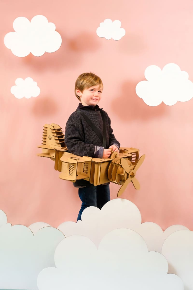 Un enfant de profil en déguisement d'avion en carton recyclé à construire, peindre et colorier soi même dans les nuages et un décor en carton sur fond rose