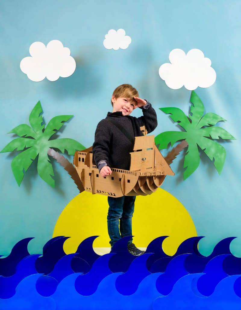 Un enfant en déguisement de bateau pirate en carton recyclé à assembler, décorer, colorier et peindre soi-même sur une île avec des palmiers et des vagues
