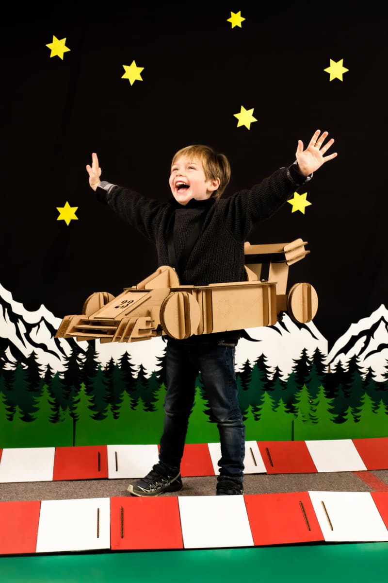 Un enfant de profil qui lève les bras en déguisement de formule 1 en carton recyclé, voiture à construire, peindre et colorier soi même sur un circuit et un décor en carton sur fond noir Tonton Carton