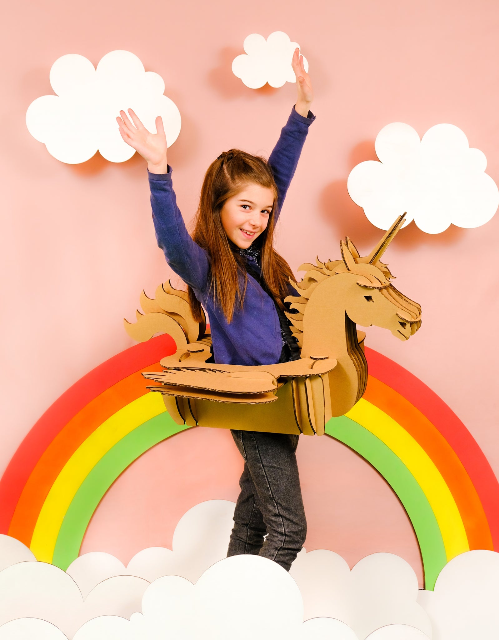 Un enfant en déguisement de licorne en carton recyclé à construire, décorer, colorier, peindre soi même devant un arc en ciel et des nuages en carton