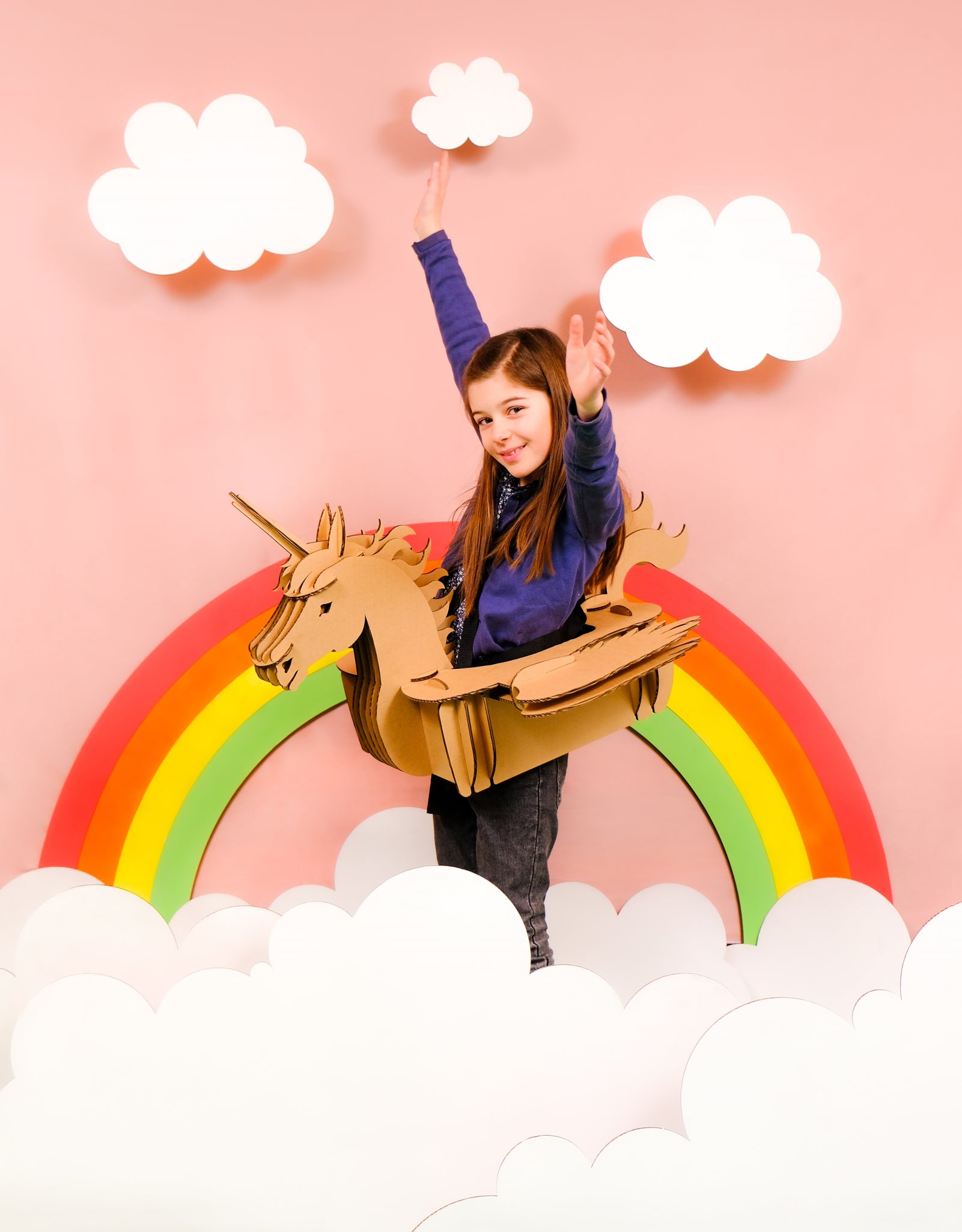 Un enfant en déguisement de licorne en carton recyclé à construire, décorer, colorier, peindre soi même devant un arc en ciel et des nuages en carton