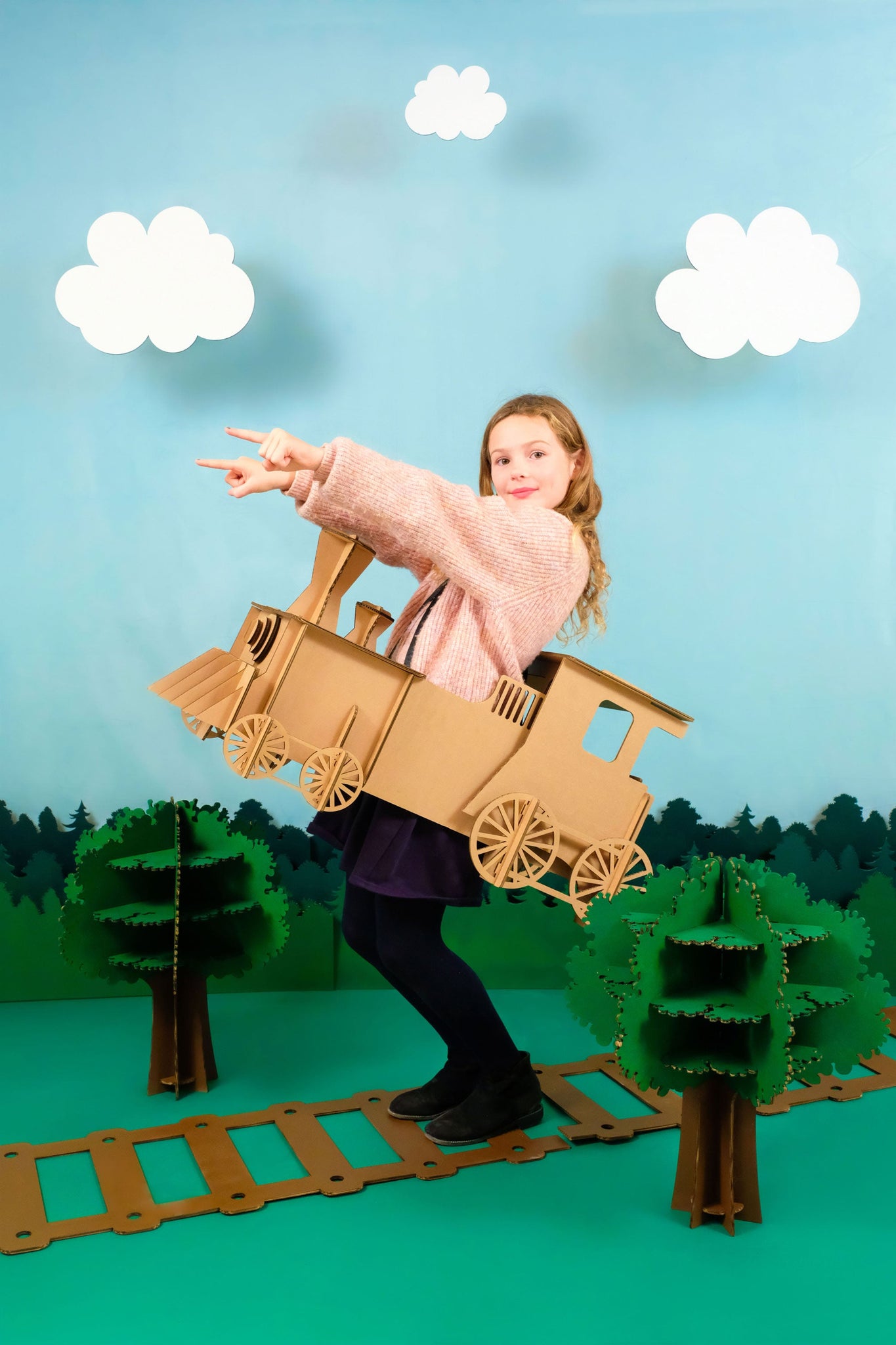 Un enfant en déguisement de train en carton recyclé à assembler, peindre et colorier soi-même sur des rails et dans une forêt avec des nuages en carton