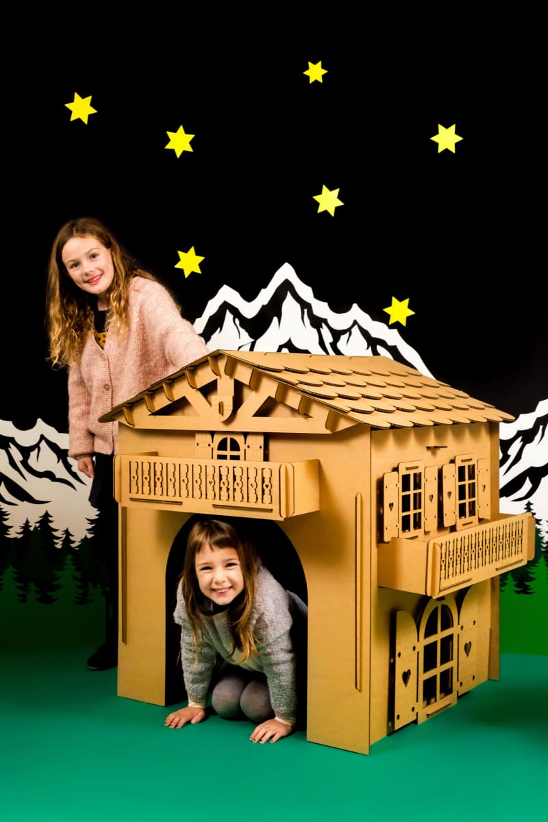 Deux enfants s'amusant dans une cabane en carton recyclé devant des montagnes et des sapins en carton, maison de type chalet savoyarden carton à construire, décorer, peindre et colorier soi même.